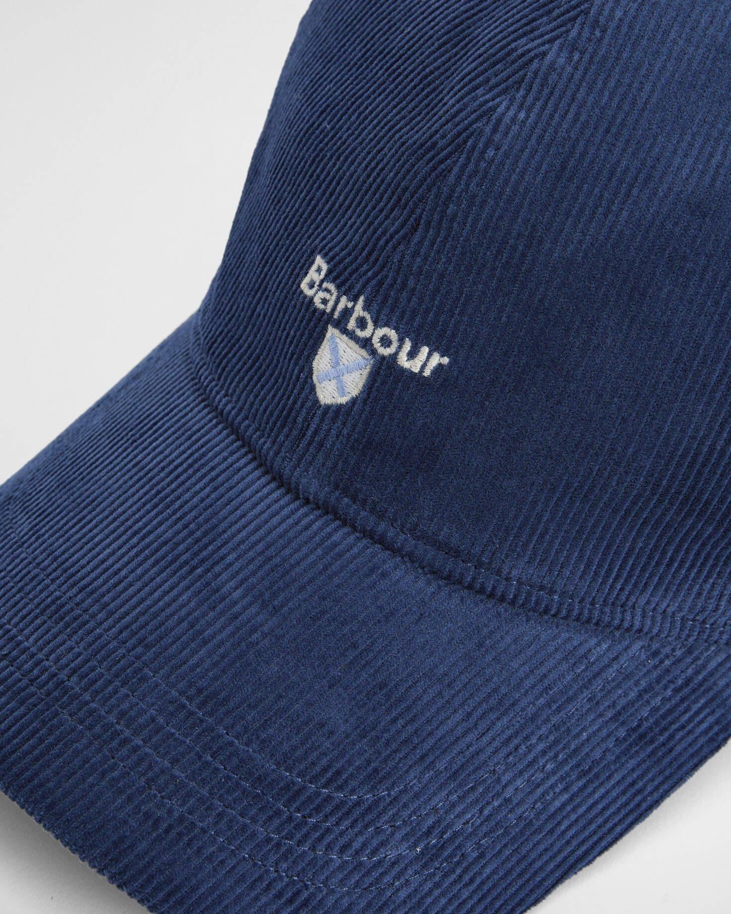 Nærbilde av Barbour caps. Ser struktur, logo med våpenskjold under. Plassering rett over brem