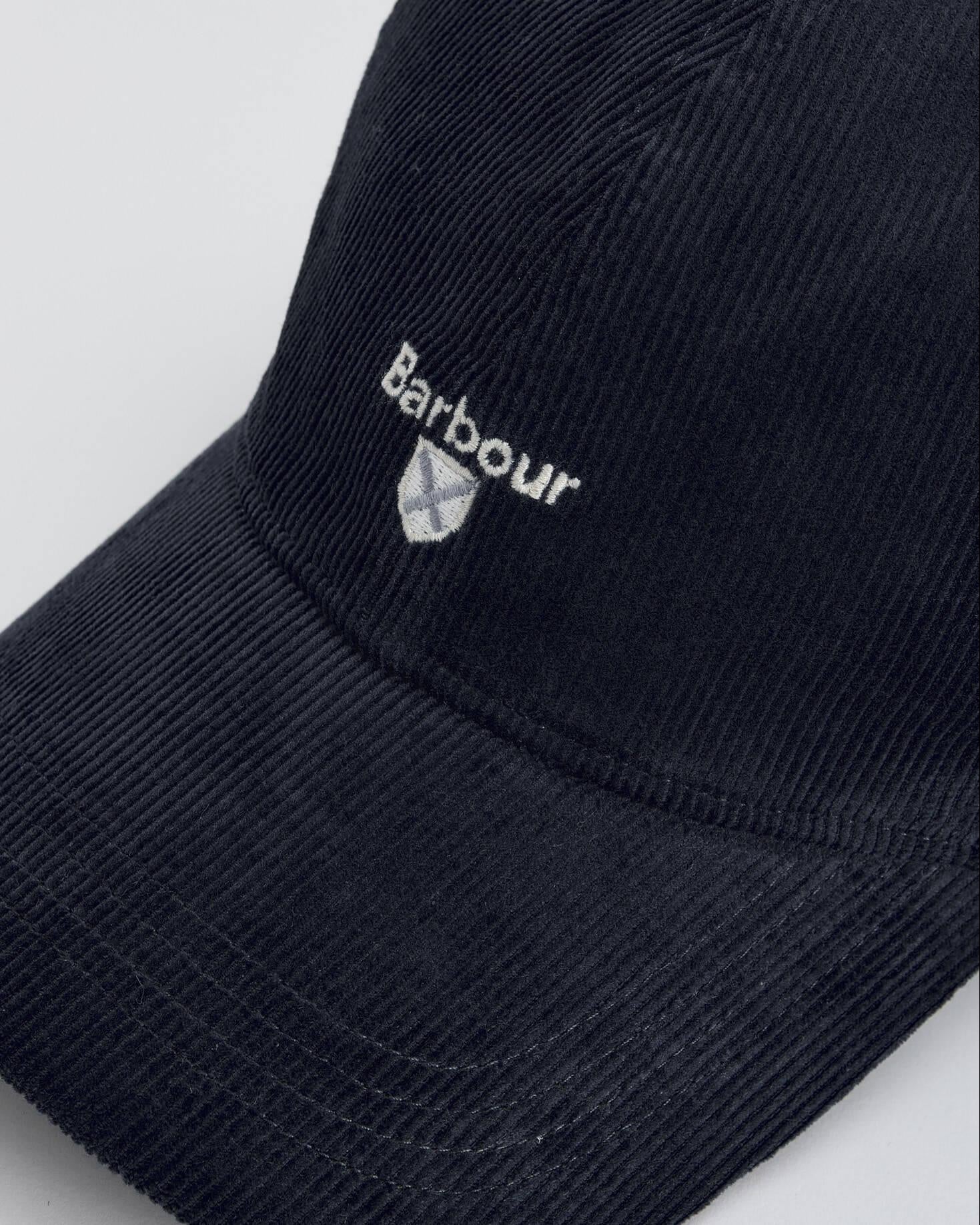 Nærbilde av Barbour caps. Ser struktur, logo med våpenskjold under. Plassering rett over brem