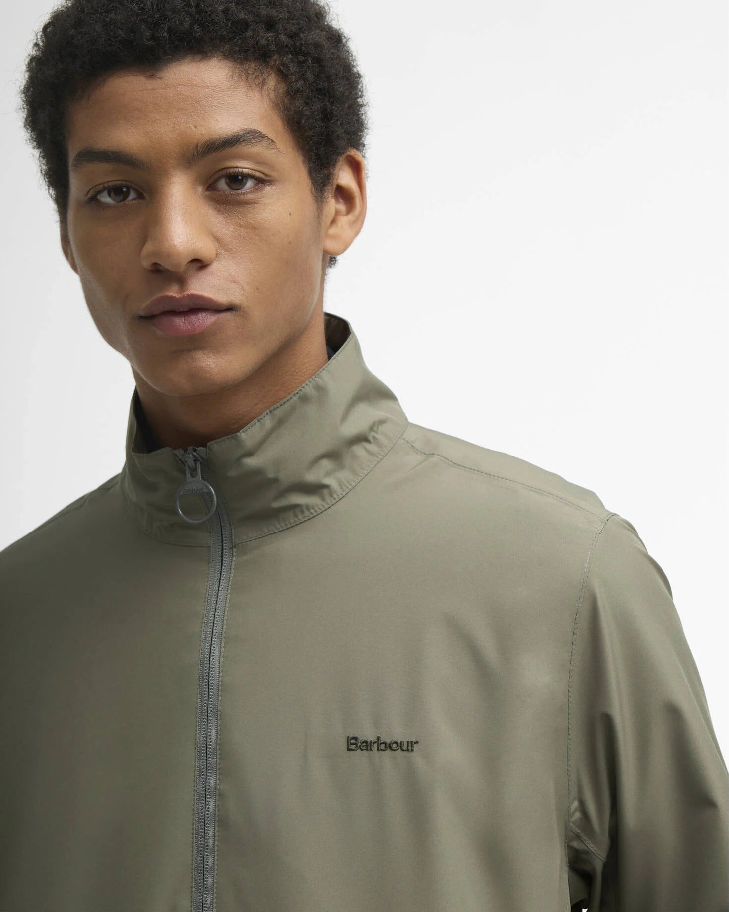 Detaljbilde av olivengrønn Barbour jakke som viser høy krage, toveis glidelås og brodert Barbour-logo på brystet.